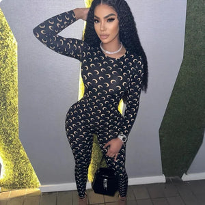 Woman wearing a black patterned outfit with a black handbag against a gray wall.
